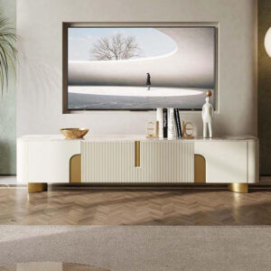 Anat Urbane luxury TV console unit featuring a long, low profile with a white/cream marble-look top, a central ribbed door/drawer panel, side cut-outs with gold backing, and low gold metal feet. Luxury furniture available near Ambala, Haryana, and across India.