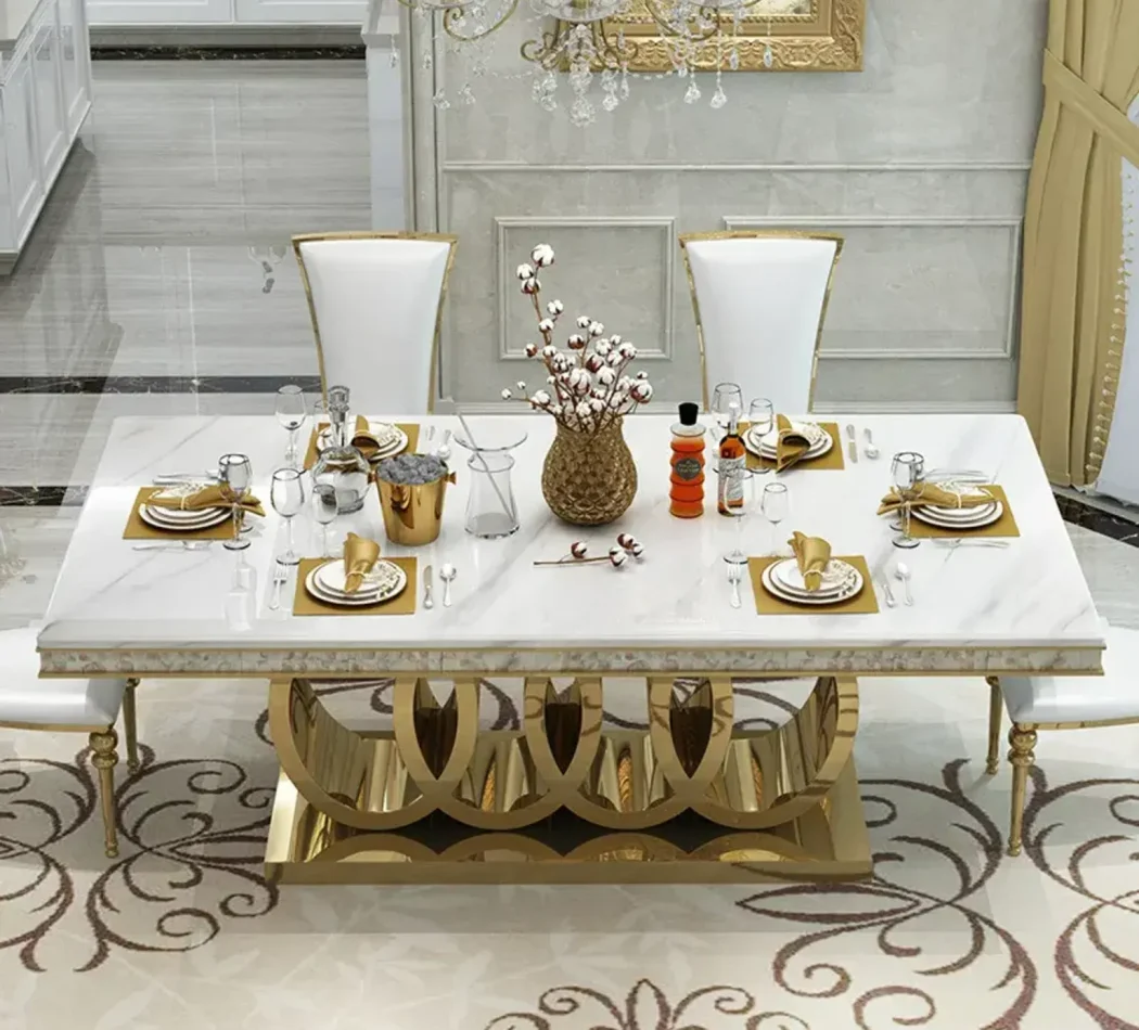 Anat Audi luxury dining set featuring a large rectangular white marble-look top, a highly decorative gold stainless steel base with a semicircular design, and high-back white upholstered dining chairs. Designer furniture available near Ambala, Haryana, and across India.